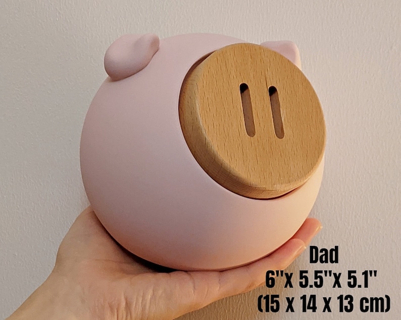 May include: A pink ceramic piggy bank with a wooden coin slot. The piggy bank is 6 inches by 5.5 inches by 5.1 inches, or 15 centimeters by 14 centimeters by 13 centimeters.