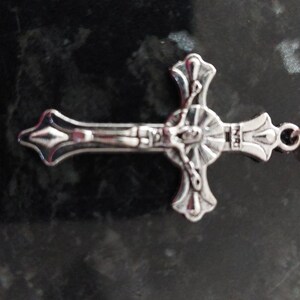 May include: A silver cross with a detailed design of a figure on the cross. The cross is attached to a white pearl bead.