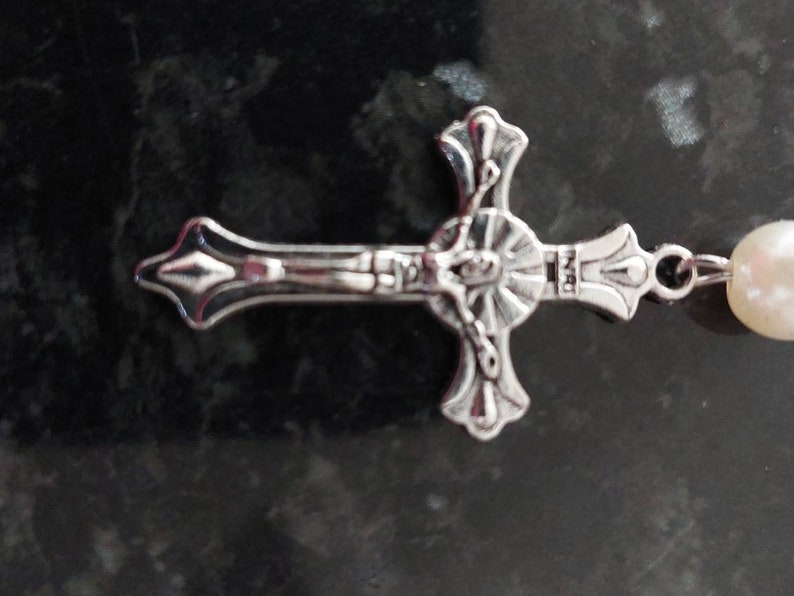 May include: A silver cross with a detailed design of a figure on the cross. The cross is attached to a white pearl bead.