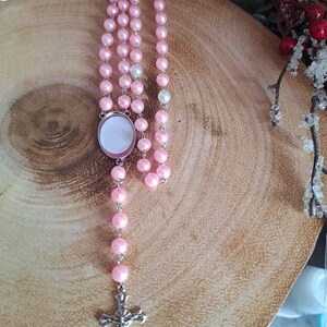 May include: A pink and white beaded rosary with a silver cross and a round, silver charm with a blank space for a picture.