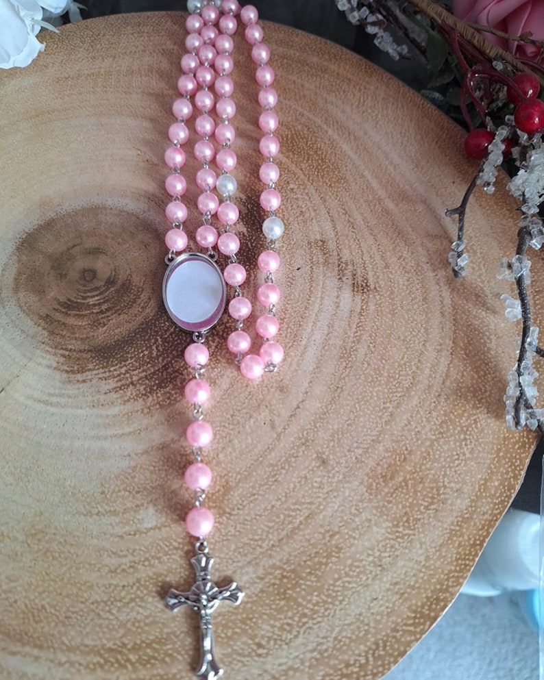 May include: A pink and white beaded rosary with a silver cross and a round, silver charm with a blank space for a picture.
