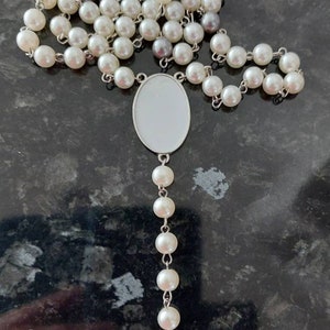 May include: A white pearl rosary with a silver crucifix and a silver oval pendant. The rosary is laid out on a dark gray surface.