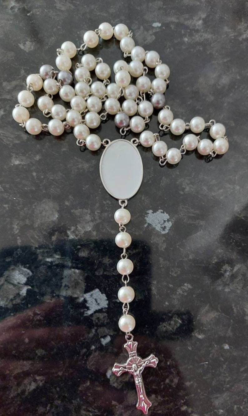 May include: A white pearl rosary with a silver crucifix and a silver oval pendant. The rosary is laid out on a dark gray surface.