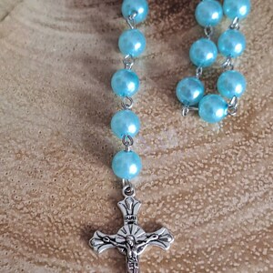 May include: A light blue rosary with a silver crucifix. The crucifix has the inscription "INRI" on the crossbar.