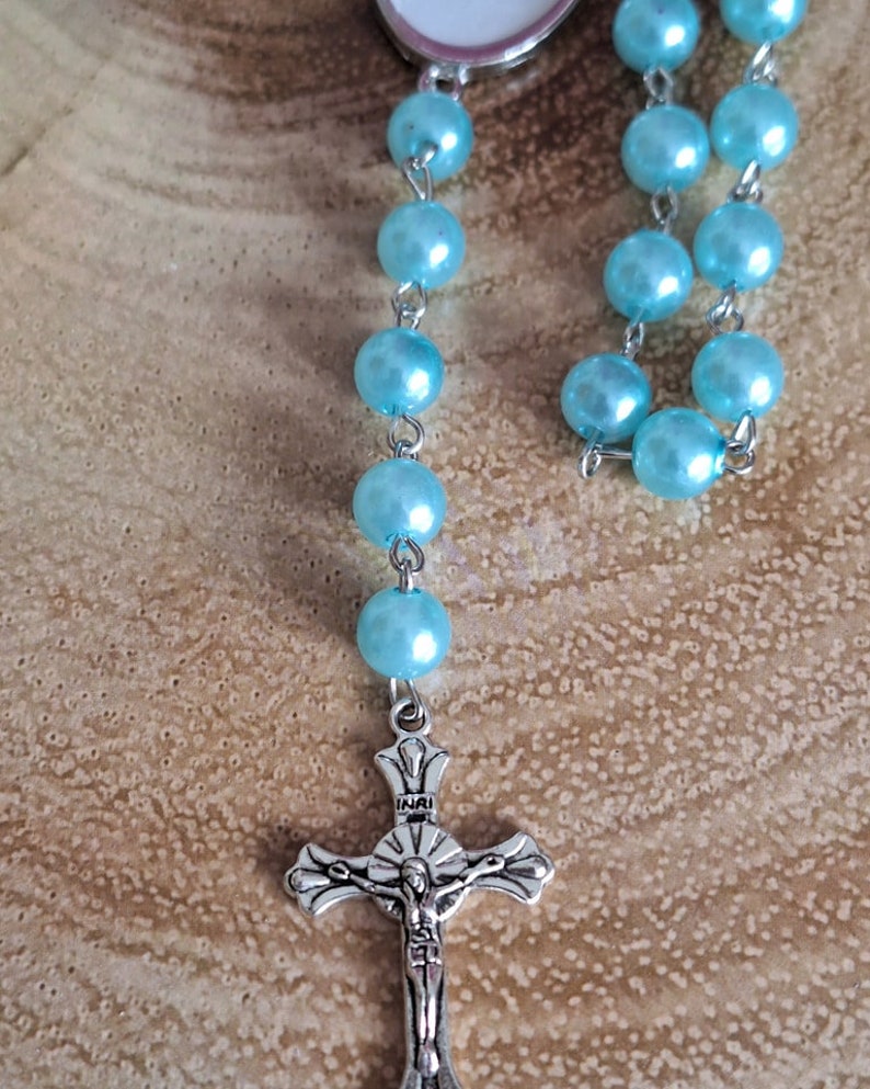 May include: A light blue rosary with a silver crucifix. The crucifix has the inscription "INRI" on the crossbar.