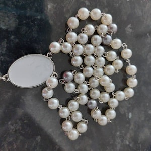 May include: A white pearl rosary with a silver oval centerpiece. The rosary is made of small, round pearls and has a silver chain.