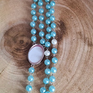 May include: A light blue beaded rosary with a silver oval charm. The charm has a blank white surface.