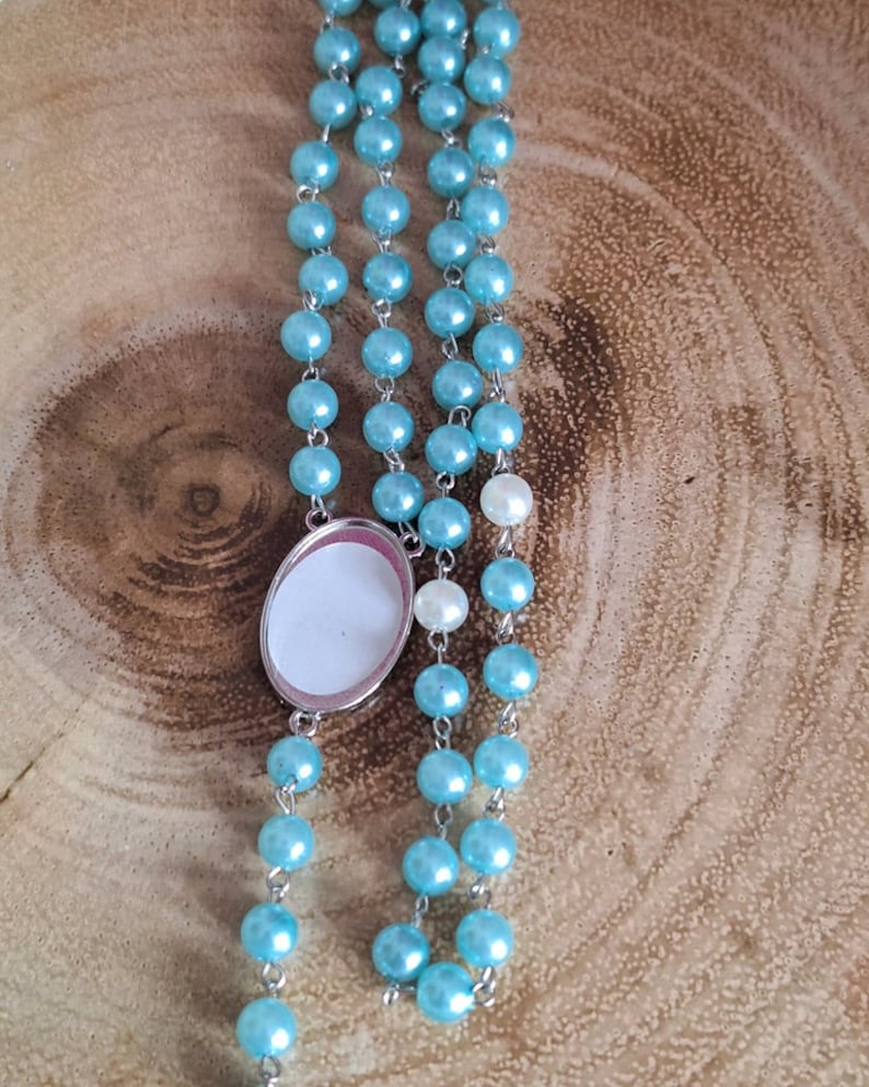 May include: A light blue beaded rosary with a silver oval charm. The charm has a blank white surface.