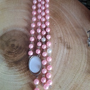 May include: A pink and white pearl rosary with a silver oval charm. The rosary is made of small, round beads and is strung on a silver chain.
