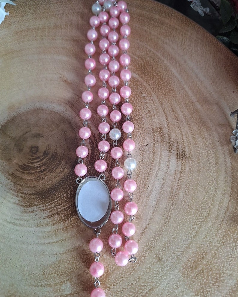 May include: A pink and white pearl rosary with a silver oval charm. The rosary is made of small, round beads and is strung on a silver chain.