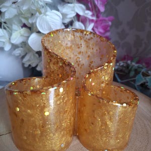 May include: A decorative, three-section candle holder. The holder is made of clear resin with gold glitter embedded throughout. The candle holder is on a wooden surface, with white and pink flowers in the background.