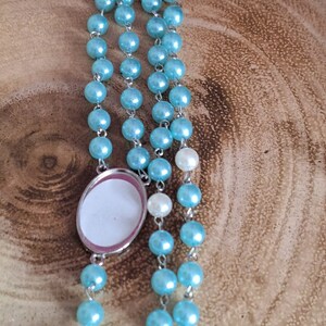 May include: A light blue rosary with a silver oval charm. The charm has a white background and a silver border.