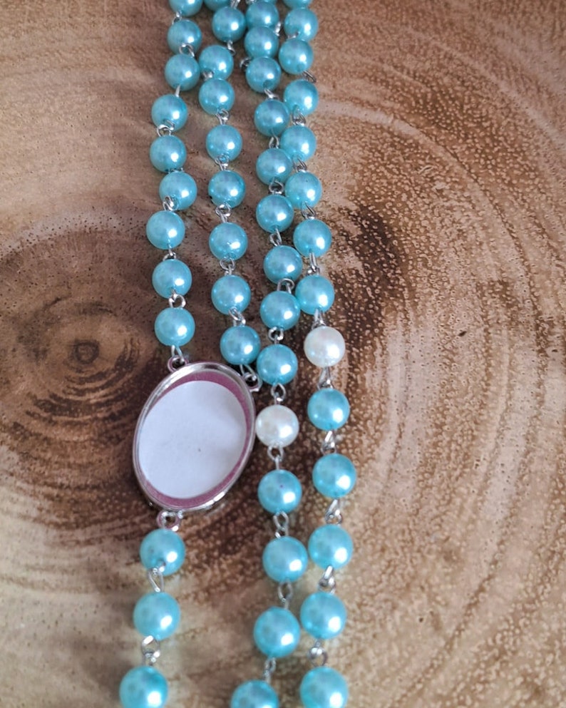 May include: A light blue rosary with a silver oval charm. The charm has a white background and a silver border.