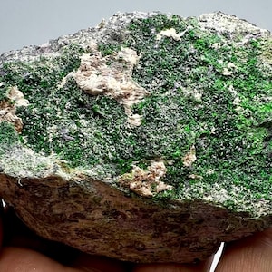 Op de afbeelding: Een ruwe, grijze rots met een grote vlek van felgroene kristallen. De kristallen zijn geclusterd en hebben een glanzende, metalen uitstraling.