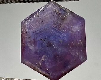 May include: A faceted, hexagonal gemstone with a deep purple hue and a central blue core. The gem is held by a pair of silver tweezers, showcasing its geometric design and crystalline structure. The stone's facets catch the light, revealing its internal patterns.