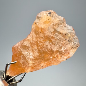 Peut inclure: Une pierre précieuse brute de couleur orange avec un aspect translucide. La pierre a une forme naturelle et irrégulière et est maintenue par un outil métallique. De petits points noirs sont dispersés sur la surface.