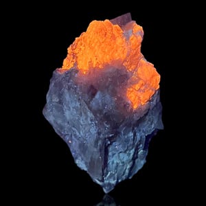 Könnte beinhalten: Ein rohes Mineralexemplar mit einer gezackten, kristallinen Struktur. Der obere Teil leuchtet in einer lebendigen, intensiven orangefarbenen Fluoreszenz, während der untere Bereich einen kühlen, gedämpften blaugrauen Ton vor einem soliden schwarzen Hintergrund aufweist. Die Buchstaben LW sind in der oberen linken Ecke sichtbar.