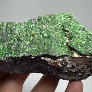 Op de afbeelding: Een ruwe rots met een levendig groene, mosachtige oppervlakte. De basis van de rots is donker, houtskoolkleurig, met witte en beige vlekken. De textuur lijkt ruw en ongelijkmatig, met natuurlijke minerale formaties. De rots wordt in een hand gehouden.