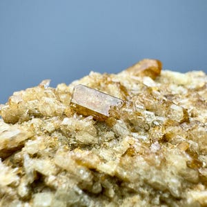 May include: Close-up of a mineral specimen featuring a cluster of light brown, crystalline structures. The crystals have a translucent quality, with a larger, rectangular crystal prominently displayed. The background is a soft, muted blue.
