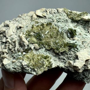 Puede incluir: Un espécimen mineral texturizado con una mezcla de colores. La base es un gris verdoso moteado, con parches de beige claro y grupos de cristales verdes translúcidos vibrantes. La forma general es irregular y áspera.