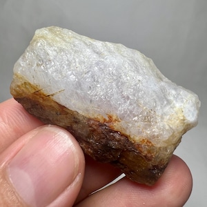 May include: A rough, natural crystal formation with a translucent, off-white upper portion and a darker, brown-toned base. The crystal is held in a hand, showcasing its raw, unpolished texture and natural color variations.