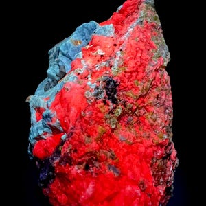 May include: A vibrant, textured mineral specimen with a striking red and blue color palette. The rock formation has a rough, uneven surface, with areas of bright red and patches of blue-gray. The letters "M.W" are in the top left corner.