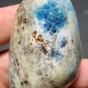May include: A polished, oval-shaped stone with a mottled appearance. The stone features areas of light blue, white, and tan, with a striking patch of deep blue veining. The surface has a natural, organic texture.