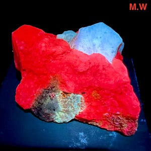 May include: A vibrant, glowing red mineral specimen with a light blue crystal formation. The rock has a rough, textured surface with areas of gray and green. The letters "M.W" are in the top right corner.