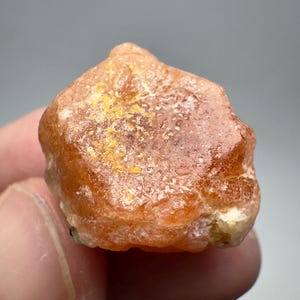 May include: A rough, orange-brown gemstone with a crystalline structure. The surface has a slightly rough texture with patches of yellow. The stone is held in a hand, with a neutral background.
