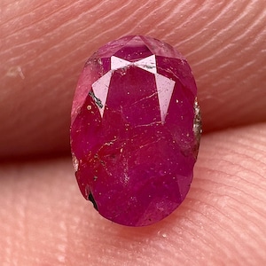 May include: An oval-cut, deep magenta-red ruby gemstone featuring a faceted surface with visible natural inclusions and internal fractures, displaying a translucent quality and a polished finish across its top surface.