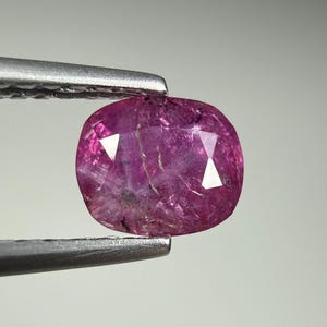May include: A cushion-cut gemstone with a vibrant magenta-pink hue and visible internal inclusions. The stone features multiple facets that reflect light, creating a sparkling appearance. It is held securely by a pair of metal tweezers.