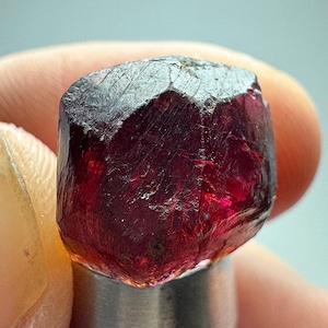 May include: A deep red, translucent gemstone with a faceted, multi-sided structure. The crystal displays a rich crimson hue with visible internal inclusions and sharp, angular edges, reflecting light across its polished, irregular surfaces.