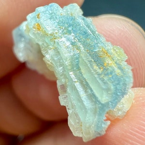 Può includere: Un gruppo di cristalli traslucidi di colore blu chiaro con una texture stratificata e striata. I cristalli hanno una leggera lucentezza iridescente e sono impreziositi da piccole inclusioni dorate.