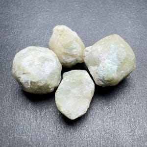 Pode incluir: Quatro pedras preciosas brutas e não cortadas, em tons de verde pálido e branco. As pedras têm uma superfície rugosa e texturizada e formas variadas, com um acabamento mate. As pedras estão dispostas sobre uma superfície cinzenta escura.