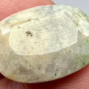 May include: A white oval gemstone with green and gray inclusions. The gemstone has a smooth, polished surface.