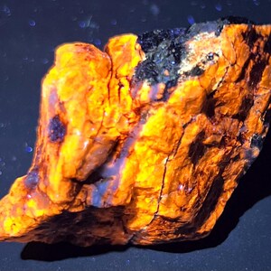 May include: A close-up of a rough, irregularly shaped rock with a vibrant orange and yellow color. Darker patches of black and blue are visible, adding contrast. The rock's surface has a textured, cracked appearance, suggesting a natural mineral specimen.