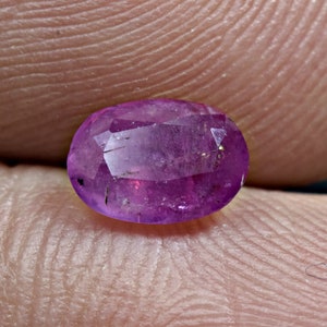 May include: A vibrant purple oval-shaped gemstone with a faceted cut. The gemstone has a slight shimmer and is set against a neutral background.