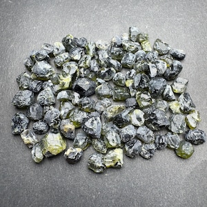 May include: A pile of rough-cut, natural peridot gemstones. The stones vary in size and shape, with a mix of dark green and olive-green hues. The gemstones are scattered across a dark gray surface, showcasing their natural, unpolished appearance.