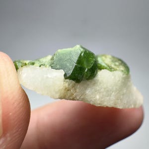 Puede incluir: Primer plano de una formación de cristales verdes sobre una base mineral blanca y verde. El cristal es de color verde esmeralda intenso con facetas geométricas y afiladas. La base mineral es de color blanco roto claro.