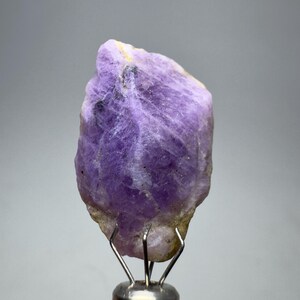May include: A raw, uncut amethyst crystal with a deep purple hue. The gemstone is mounted on a silver metal stand, showcasing its natural, textured surface and crystalline structure. The amethyst is a vibrant, natural mineral.