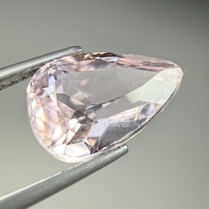 May include: A pear-shaped, light pink gemstone with multiple facets that reflect light. The translucent stone is held by a pair of silver-toned metal tweezers, showcasing its clear, pale pink color and precise, sharp-edged cut against a neutral background.