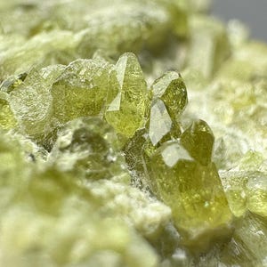 Könnte beinhalten: Nahaufnahme eines Clusters hellgrüner Kristalle mit scharfen, facettierten Kanten. Die Kristalle sind vor einem Hintergrund aus einer helleren, strukturierten mineralischen Basis angeordnet, wodurch ein natürliches, erdiges Aussehen entsteht. Die Kristalle sind durchscheinend.