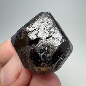 May include: A dark, faceted mineral specimen with a deep black color and a reflective, crystalline surface. The rough-hewn edges and natural texture of the stone are visible. The mineral is held in a hand against a neutral background.