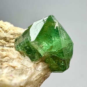 Pode incluir: Uma vibrante formação de cristal verde com facetas geométricas nítidas. O cristal está sobre uma base mineral bege áspera. A clareza e a cor da gema são impressionantes, sugerindo um espécime de alta qualidade.
