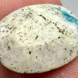 May include: A white oval cabochon gemstone with black and blue inclusions. The gemstone has a rough, textured surface.