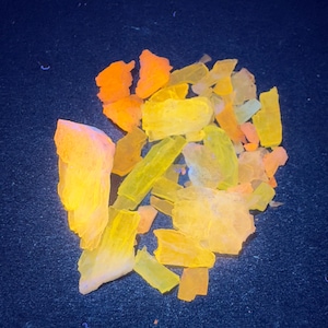 May include: A collection of irregularly shaped, translucent crystals in shades of yellow, orange, and pale pink. The crystals are scattered on a dark blue surface, creating a vibrant and eye-catching display. The crystals appear to be illuminated from within.
