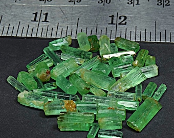 Op de afbeelding: Een verzameling groene smaragd kristallen, sommige met een lichte bruine tint, zijn gerangschikt op een zwart oppervlak. Een liniaal is zichtbaar op de achtergrond, wat aangeeft dat de kristallen ongeveer 2,5 cm lang zijn.