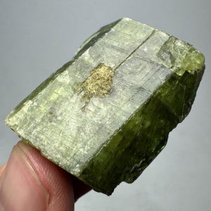 May include: A raw, green mineral specimen with a crystalline structure. The stone has a light green hue with darker green edges and a gold-colored inclusion. The mineral is held in a hand against a neutral background.