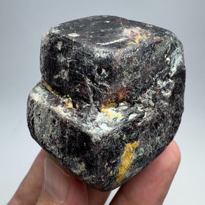 Pode incluir: Um espécime mineral escuro e retangular com uma superfície áspera e texturizada. A pedra é principalmente marrom escuro com áreas vermelhas, brancas e amarelas. O mineral é segurado em uma mão contra um fundo claro.