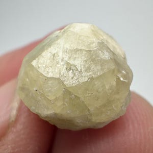 Pode incluir: Uma gema facetada amarelo claro com uma estrutura cristalina. A gema tem uma textura ligeiramente áspera e uma aparência translúcida. A pedra é segurada pelos dedos de uma pessoa.
